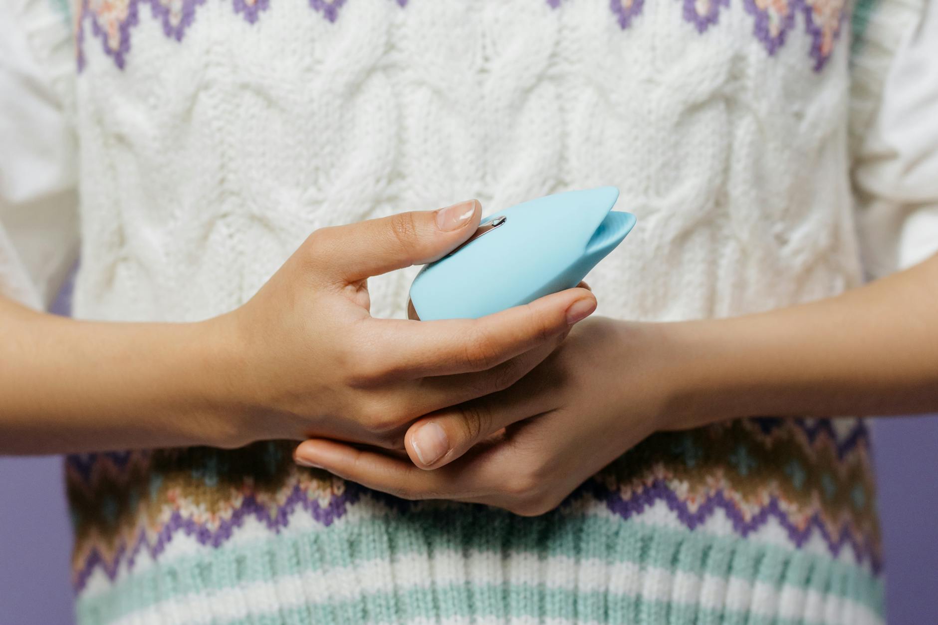 Hands holding a blue personal massager against a knitted sweater, representing solo pleasure and self-awareness.